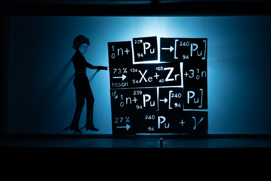 This is a silhouette, shadow puppet image, it is black and white with hints of blue in the shadows. A shadow puppet figure of a woman is standing next to a large block of tetris like shapes piled together, the shapes show chemical symbols and equations including plutonium, Xenon and Zirconium.