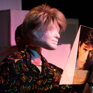 A close up of Alan looking at a Bryan Ferry record. Alan is a full sized male puppet with an 80s wham style haircut, he is wearing a vibrant patterned shirt. In the background we can see some white blocks out of focus.