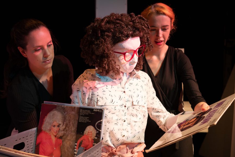 Kate, a female human sized puppet with red 80s glasses and a tight perm, is standing in a record shop looking through records. There is a box of vinyl on a desk, one record is visible showing Dolly Parton in a red shirt with very big 80s hair. There are also two puppeteers partially visible in the image, they are each holding one of Kate’s hands.