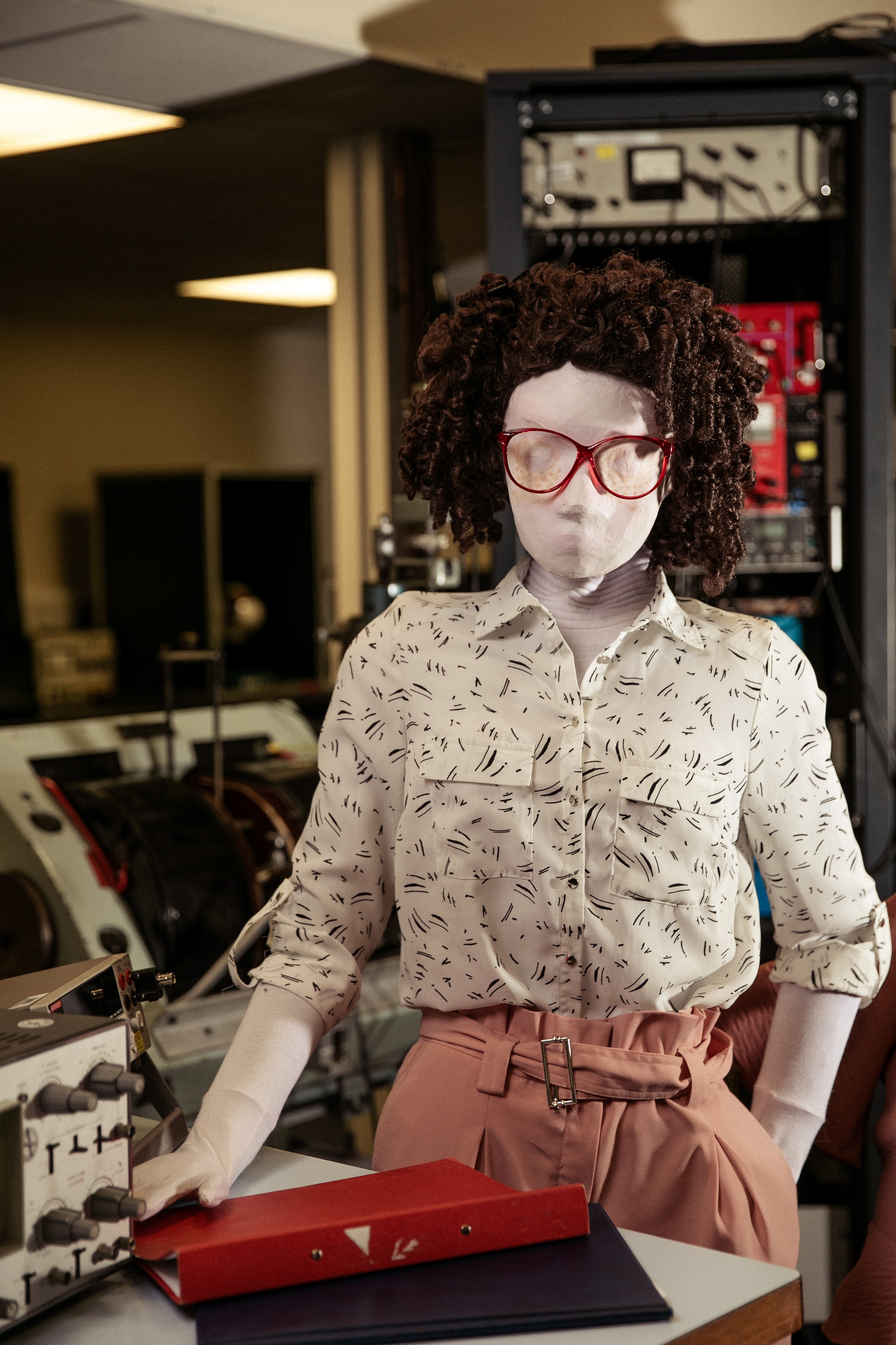 Kate, a female human sized puppet with big, red, 80s glasses and a tight perm, stands behind behind a desk with one hand resting on her red folder, and one hand in her pocket. There is an oscilloscope partially in view on the desk, with lots of physics equipment in the background.