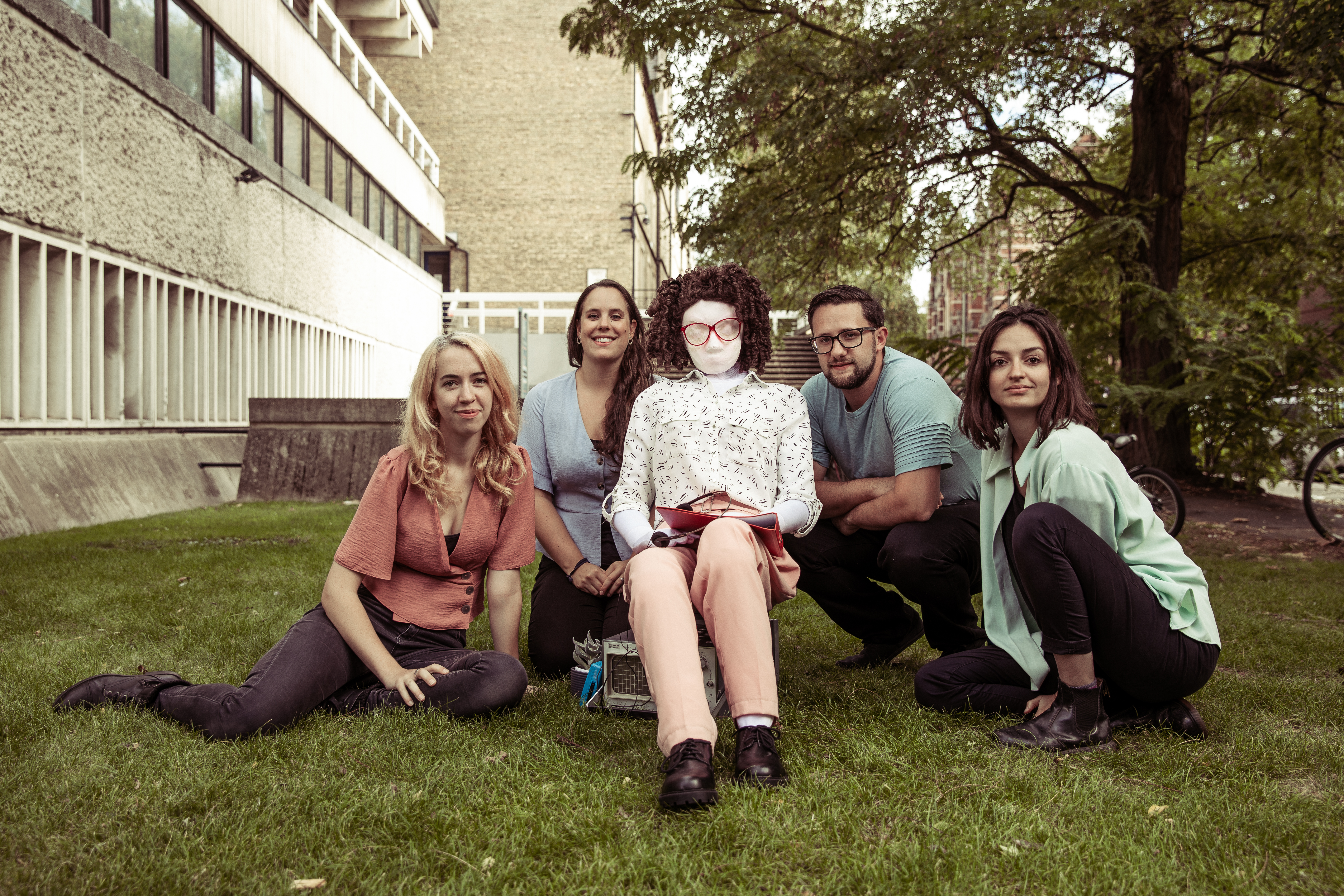 The full cast of Flux sits on the grass outside a grey, concrete building. On the far left is Hattie, with blonde hair and peach top, then Molly with long brown hair and a pastel blue top, full sized puppet Kate sits in the middle, with big 80s glasses and a tight perm. Next to Kate is Matt with short brown hair and a light blue top and he is next to Anne with shoulder length brown hair and a light green shirt.