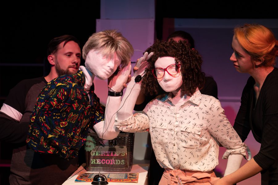 Kate, a female human sized puppet with red 80s glasses and a tight perm, is standing next to Alan, a full sized male puppet with an 80s wham style haircut. They are in a record shop represented by a box of vinyl on a desk with a sign saying Gertie’s Records. There is also a record on display and a silver shop bell. Kate and Alan are holding wire frame headphones, one side in each hand and leaning into listen to the same record. Also visible are three puppeteers, 2 holding Kate and 1 holding Alan.
