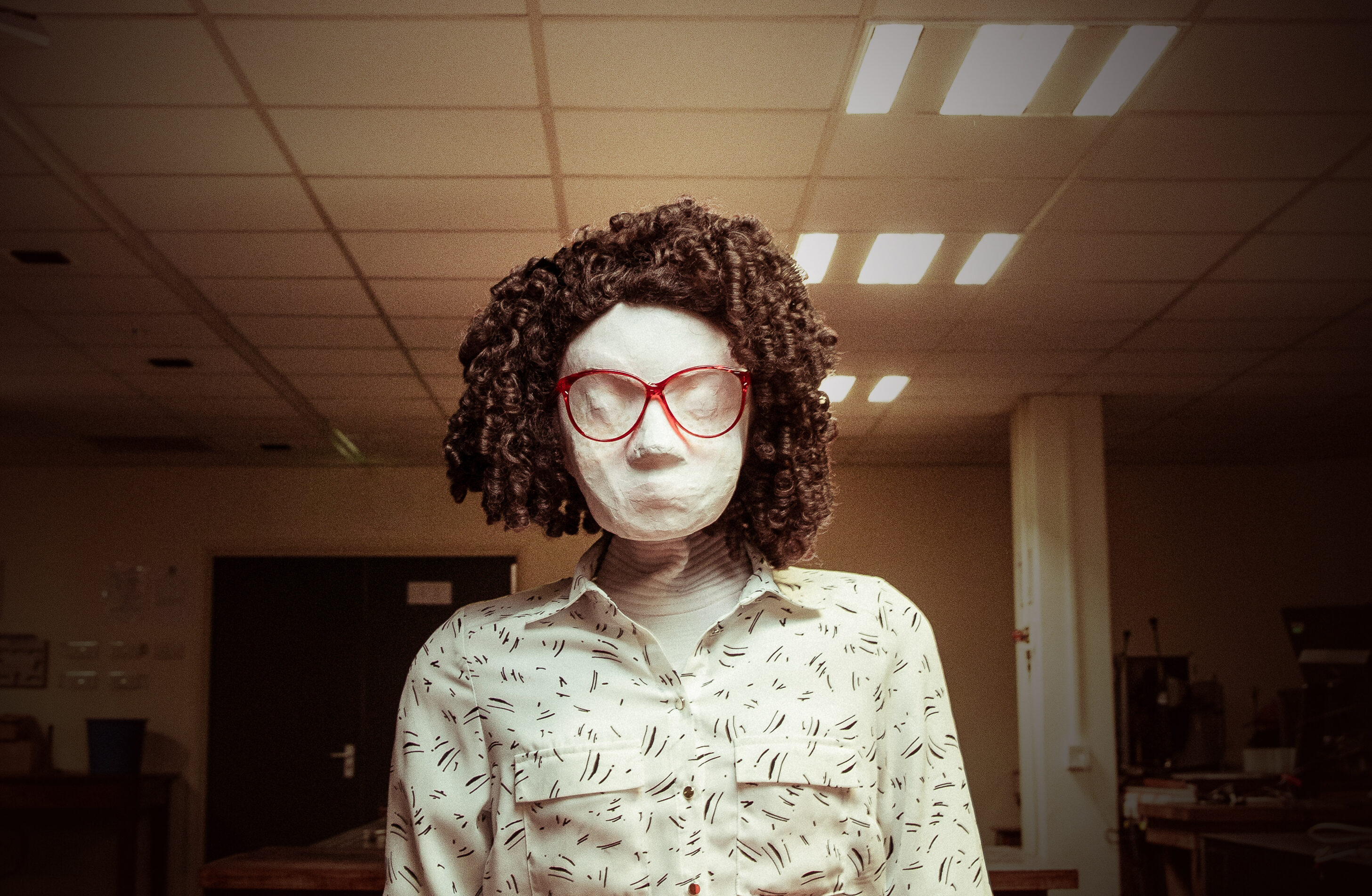 Kate, a female human sized puppet with red 80s glasses and a tight perm, sits behind a dark brown wooden desk in a badly lit office space. In the background there are some computers and equipment on other desks. Kate is looking directly at the camera.