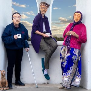 3 masked actors representing varied elderly demographics across the UK are leaning against a white geometric structure, holding cups in their hands. One actor holds a crutch, and a cat puppet is sat on the floor looking up towards them. They all look warmly towards the camera. In the background is a blue sky with clouds.