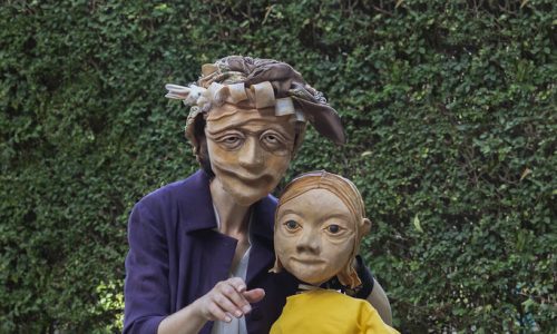 Ida, a masked character wearing a purple coat and silk scarf tied around her head, sits on a park bench with Sophie the puppet. Sophie, wearing a bright yellow jumper, is sat leaning next to her. Ida is pointing toward the camera and both are looking forward.