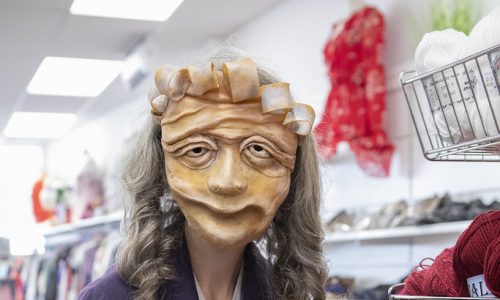 Ida, a masked character wearing a purple coat and holding 4 purple balls of wool, stands in a haberdashery and gestures one ball of wool toward the camera.