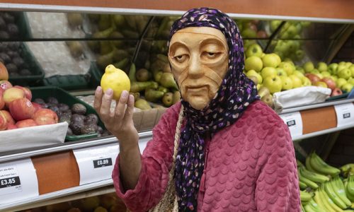 Jaanvi, a masked character wearing a headscarf, pink cardigan and blue patterned sari, stands in the fruit section of a grocery shop holding a lemon and looking toward the camera.