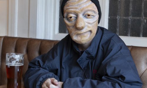 Ralph, a masked character wearing a dark blue rain jacket and black woolly hat, sits in a pub looking toward the camera. On the wooden table are a pint of beer and a metal cigarette tin.