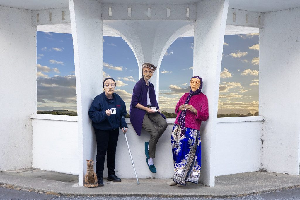 3 masked actors representing varied elderly demographics across the UK are leaning against a white geometric structure, holding cups in their hands. One actor holds a crutch, and a cat puppet is sat on the floor looking up towards them. They all look warmly towards the camera. In the background is a blue sky with clouds.
