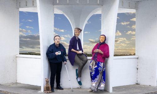 3 masked actors representing varied elderly demographics across the UK are leaning against a white geometric structure, holding cups in their hands. One actor holds a crutch, and a cat puppet is sat on the floor looking up towards them. They all look warmly towards the camera. In the background is a blue sky with clouds.