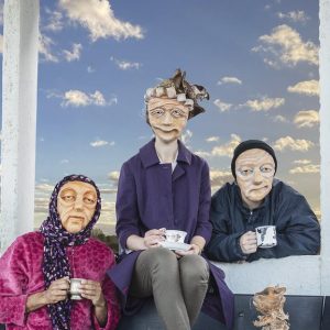 3 masked actors representing varied elderly demographics across the UK are gathered around a bench, holding cups in their hands. A cat puppet is sat on the bench looking up towards them. They all look warmly towards the camera. In the background is a blue sky with clouds.