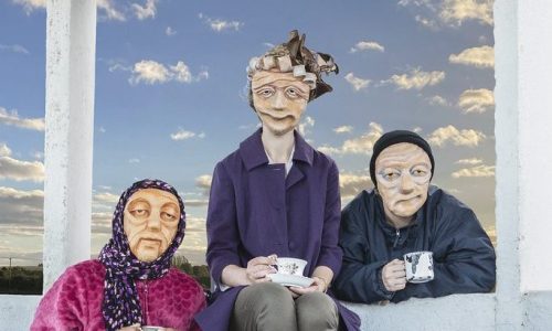3 masked actors representing varied elderly demographics across the UK are gathered around a bench, holding cups in their hands. A cat puppet is sat on the bench looking up towards them. They all look warmly towards the camera. In the background is a blue sky with clouds.
