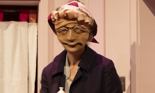 On set, Ida, a masked character, wearing a purple coat and silk scarf tied around their hair, is holding a floral tea pot in both hands and looking out toward the audience.