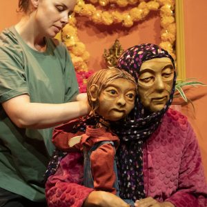 On a brightly coloured set, Anika the puppet is sitting on the lap of Jaanvi, a masked character who is wearing a purple head scarf and soft pink cardigan. They are embracing each other. Anika is being operated by another standing performer.