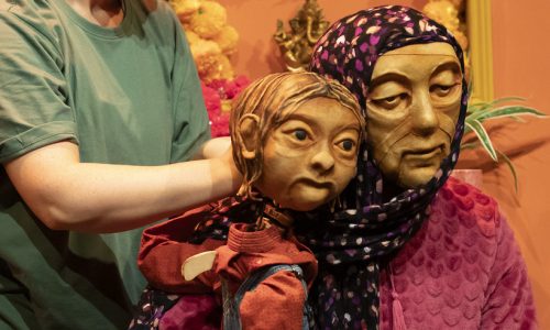 On a brightly coloured set, Anika the puppet is sitting on the lap of Jaanvi, a masked character who is wearing a purple head scarf and soft pink cardigan. They are embracing each other. Anika is being operated by another standing performer.
