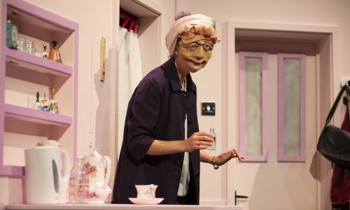 On a set with various shades of pink, Ida, a masked character, wearing a purple coat and silk scarf tied around their head, is looking out toward the audience.
