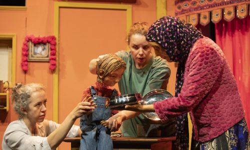 On a brightly coloured set, Anika the puppet, operated by two performers, is sat on a table looking curiously at a metal dish. Jaanvi, a masked character wearing a purple head scarf, is offering the dish to Anika.