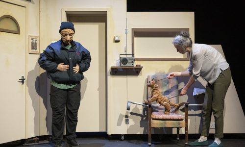 Two performers stand on a cream set. Ralph, a masked character stands facing the audience with both hands patting his jacket pockets, looking down. The other performer is leaning over a chair, operating a cat puppet, both facing toward Ralph.