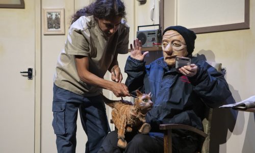On a cream set, Ralph, a masked character is sitting on an armchair, leaning back and away from a cat puppet who is perched on their lap. Another performer is leaning over the chair, operating the puppet. Ralph holds one hand up in shock, the other holds a metal cigarette box.