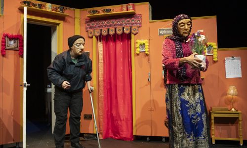 On a brightly coloured set stand two masked performers. Jaanvi, a masked character, holds a bouquet of flowers and is facing the audience. Ralph, the other masked character, stands slightly behind, using a crutch and looking toward Jaanvi.