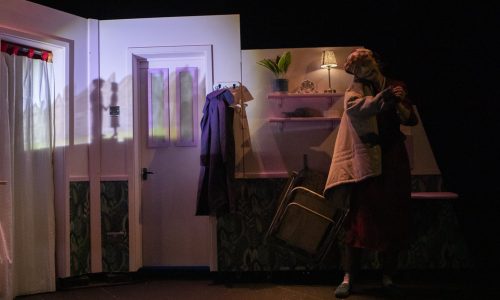 On a low lit set, Ida, a masked character is half-wearing a coat on one shoulder. She warmly leans her face onto the shoulder of the coat, holding the coat sleeve in her hand. On the back wall appear a shadow of two people facing each other and holding hands.