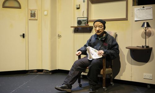 On a cream set, Ralph, a masked character sits in an armchair, holding a newspaper on his lap with one hand, and the arm rest with the other. One leg is extended as they look out toward the audience.