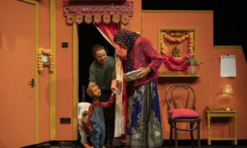 On a brightly coloured set, Anika the puppet, operated by two performers, is offering a colourful card to Jaanvi, a masked character. Jaanvi, holding a plastic tupperware box is looking warmly down toward Anika.