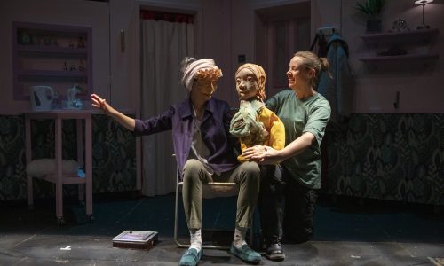 Two performers and a puppet are close together on a low lit set. Ida, a masked character, gesturing with one arm extended, is sat on a fold out chair with the puppet sat on their knee. They are looking toward each other. The other performer is smiling and crouched next to the chair, operating the puppet.