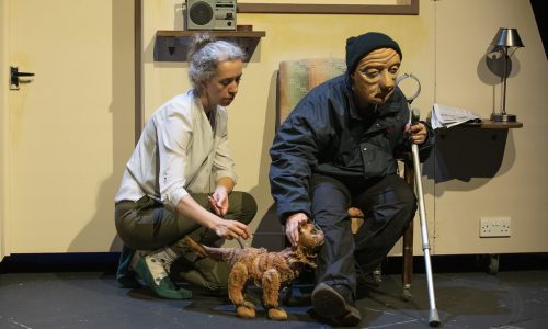 On a cream set, two performers and a cat puppet appear next to each other. Ralph, a masked character sits in an armchair, holding a crutch in one hand and leaning down to stroke the cat's head with the other. The other performer is crouched by the chair, operating the cat puppet, both facing toward Ralph.