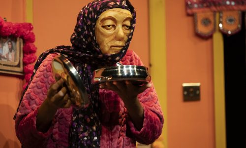 On a brightly coloured set, Jaanvi, a masked character wearing a purple head scarf, is looking out toward the audience. They are holding a metal dish close to their face and in their other hand they hold the metal lid.