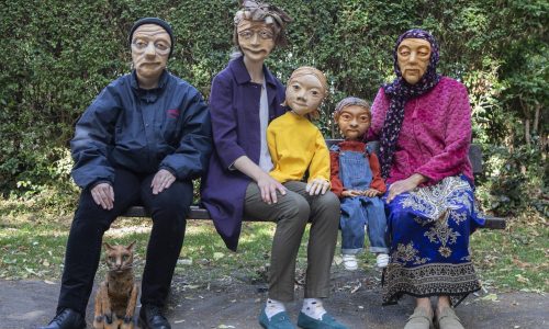 3 masked actors representing varied elderly demographics across the UK are sat on a park bench. One actor has a cat puppet sat at their feet. The other two are holding puppets. They all look warmly towards the camera.