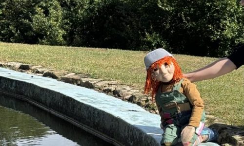 A puppet called Lyra with bright orange hair, sits at the edge of a curved stone water feature, gazing into the water. Lyra, wears a grey cap and colourful striped socks with blue shoes. Her reflection is clearly visible in the rippling water below.