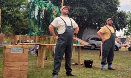 Two performers in waders, tshirts, and caps stand with hands on their hips