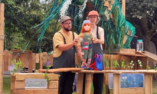 Lyra, orange-haired, in green patchwork dungarees stands beside her blue rucksack.