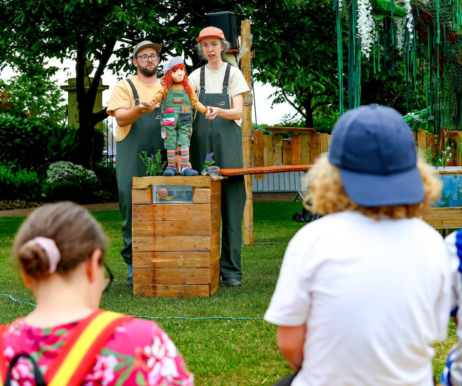 Lyra, a puppet with ginger hair wearing patchwork dungarees, stands on a wooden set talking to the audience. 